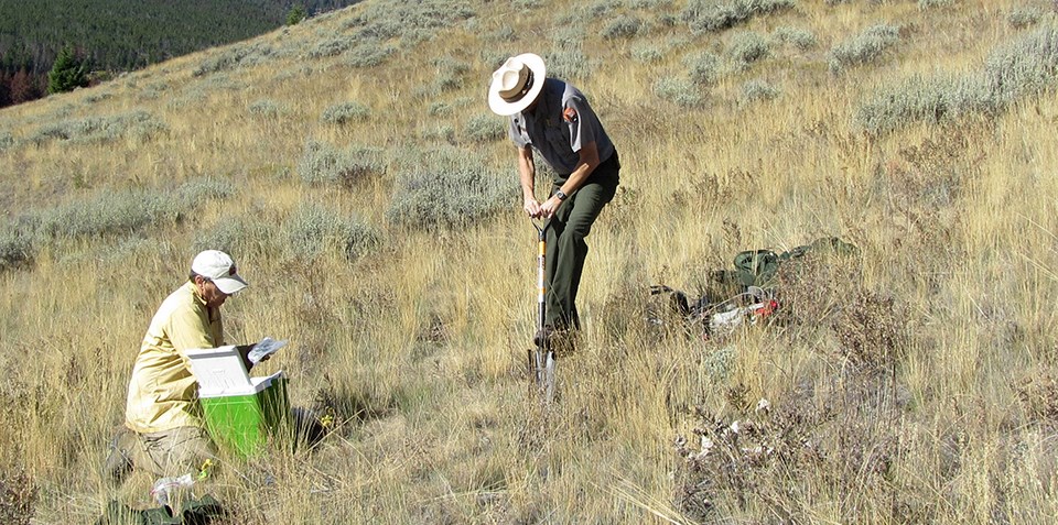 A man in NPS uniform uses a shovel on a slope while another man is nearby.