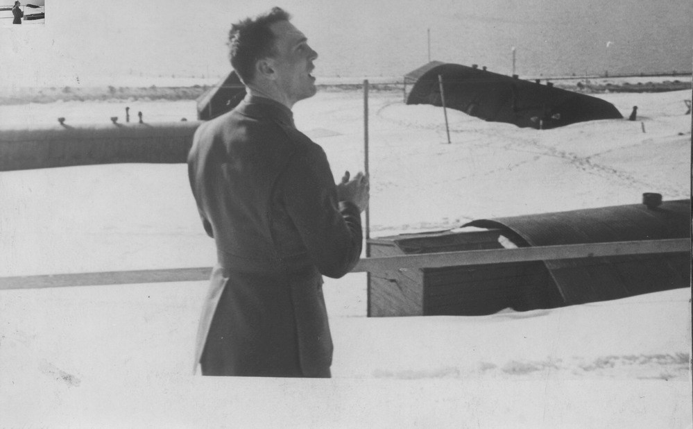black and white photo of a man in uniform, overlooking a snowy landscape with quonset huts.