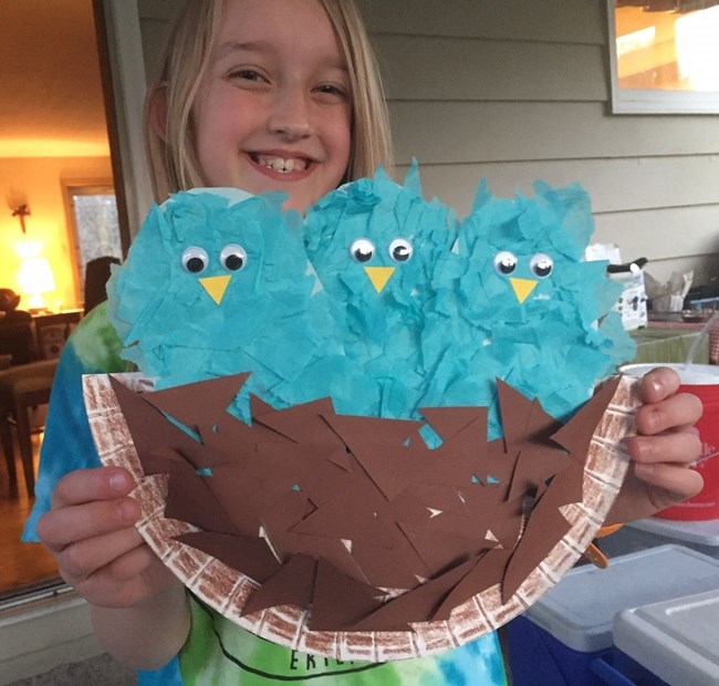 A girl holds up a paper plate craft that shows three bluebirds in a brown construction paper nest.