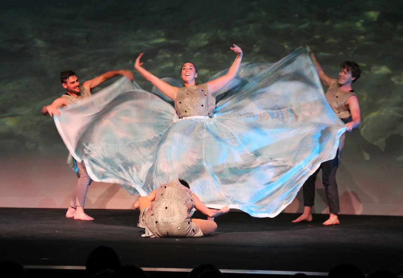 Three dancers surround woman dancer, holding her shiny, blue, parachute-like skirt.
