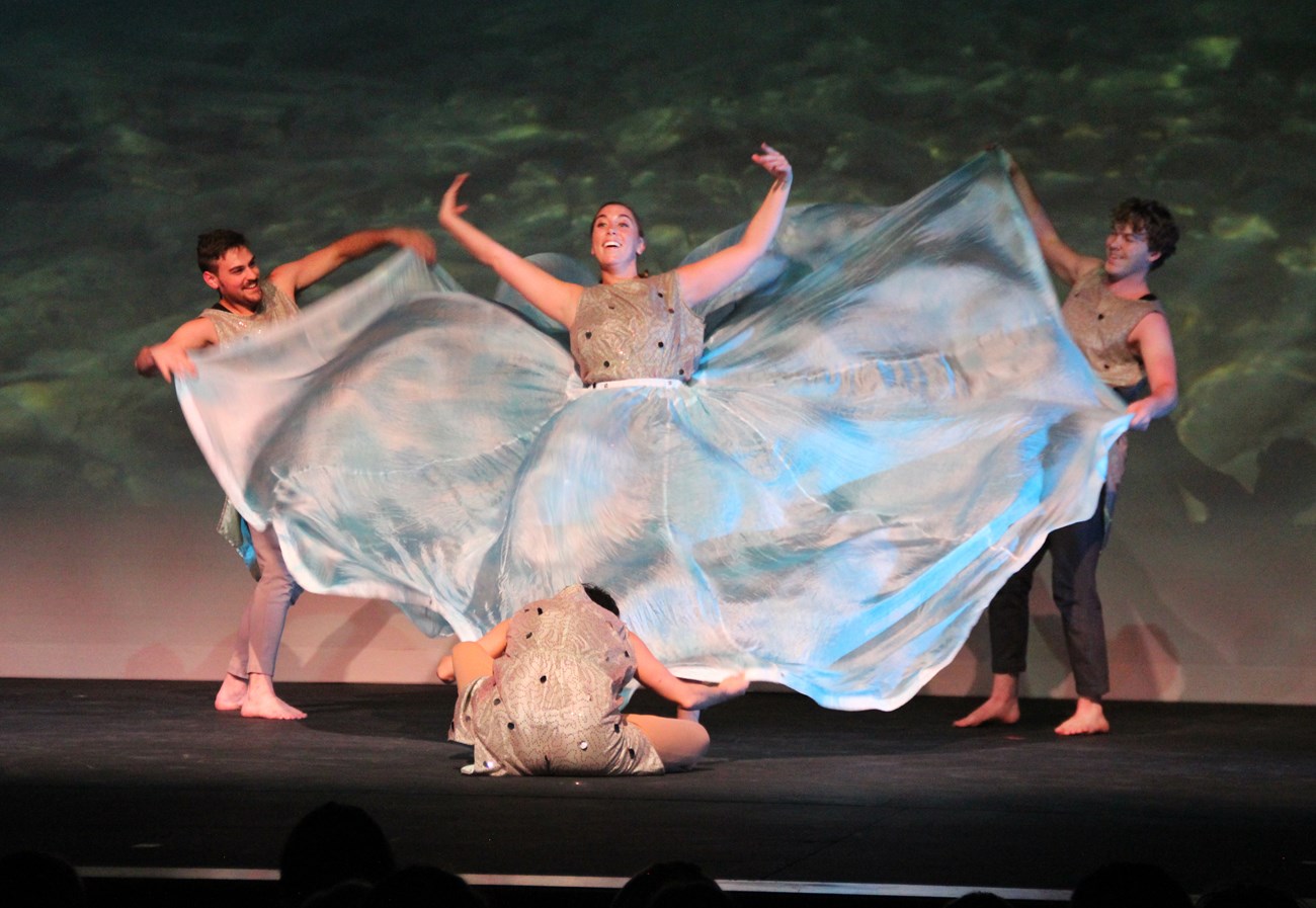 Performers dance to a song about bull trout. Three dancers surround woman dancer, holding her shiny, blue, parachute-like skirt.