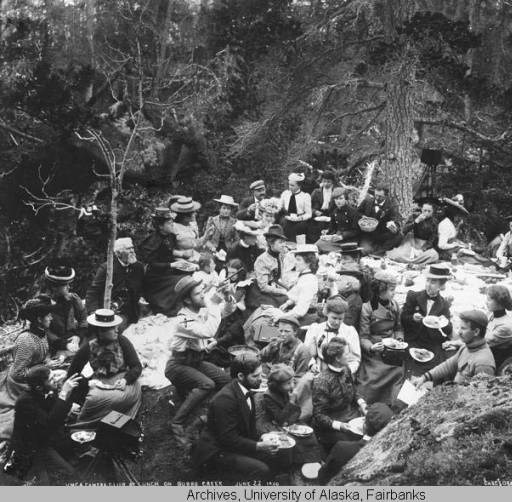Black and white image of a large group of men and women picnicking in the woods