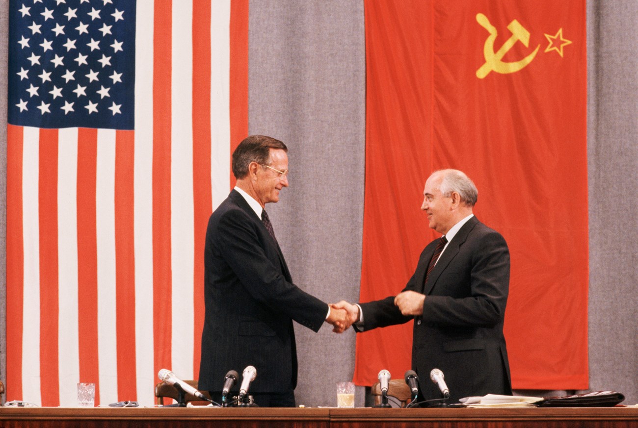 Presidents Bush and Gorbachev shake hands in front of American and Soviet Flags