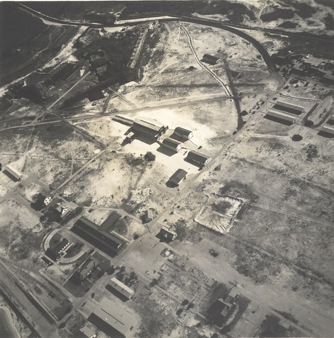 Aerial black and white photo of several buildings scattered across a sandy landscape. Aerial black and white photo of several buildings scattered across a sandy landscape.