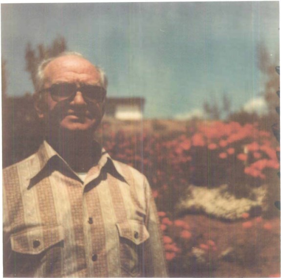 lt= Color photo of an older man posing in front of brightly colored flowers.