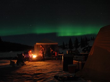 the northern lights dance over people sitting around a fire