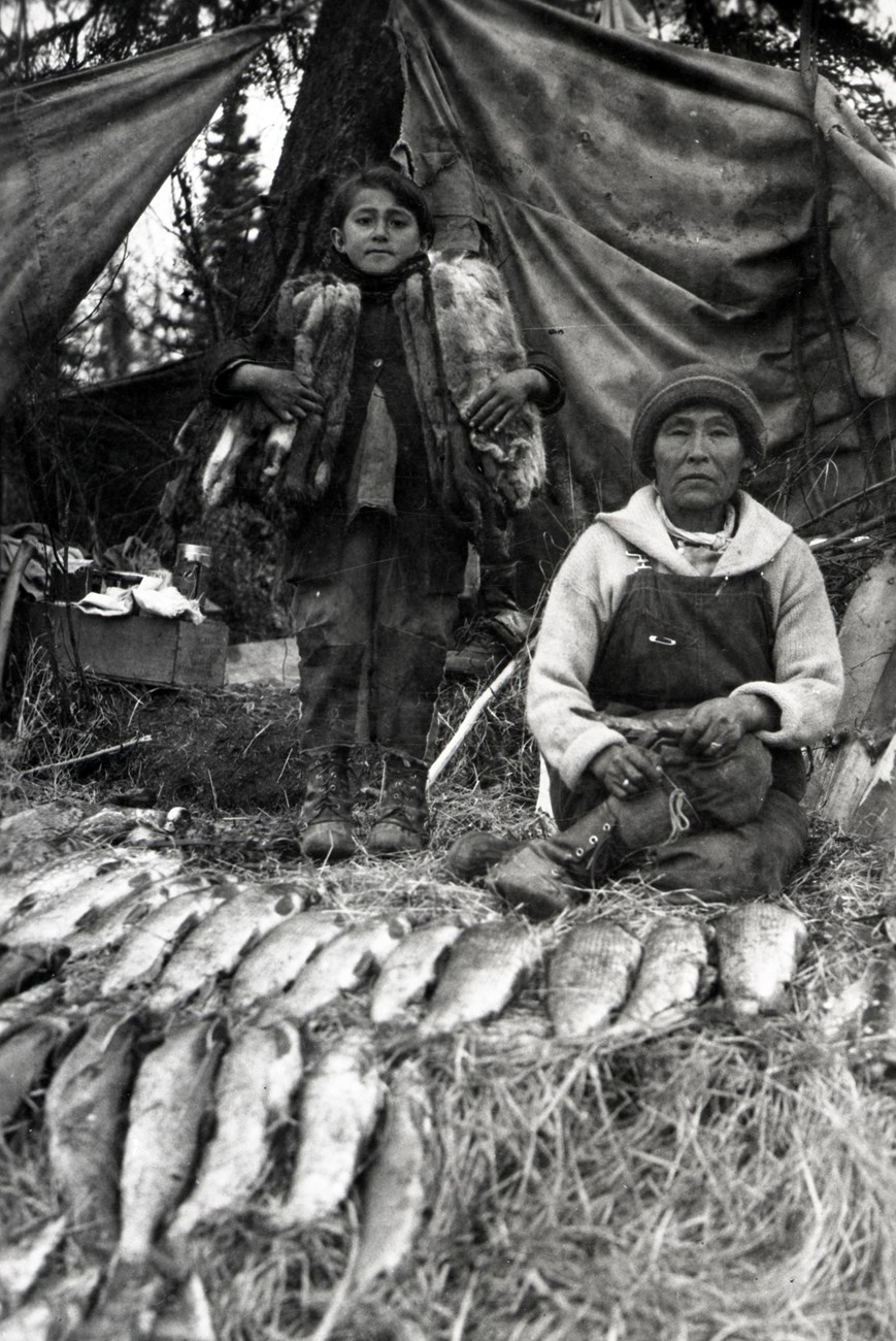Historic photo of a woman and child in front of tent with fish and furs in foreground.