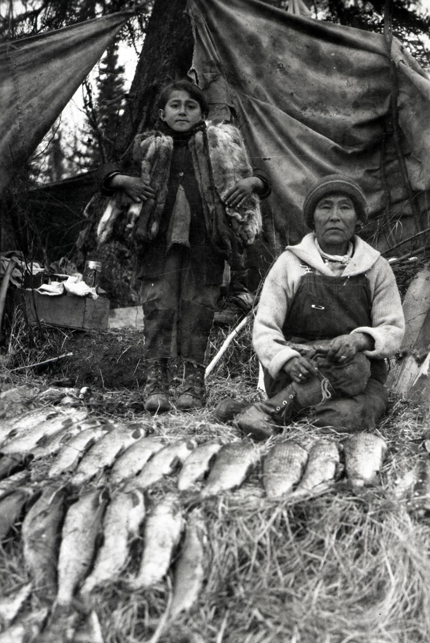 Historic photo of a woman and child in front of tent with fish and furs in foreground. Historic photo of a woman and child in front of tent with fish and furs in foreground.