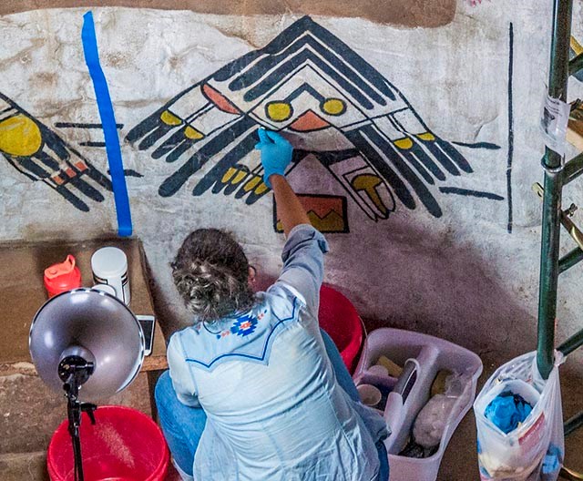 a woman wearing rubber gloves is cleaning an American Indian design that is painted onto a masonry wall.