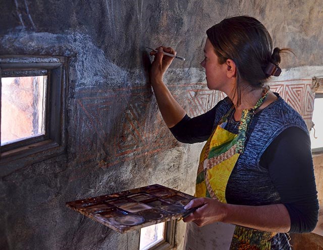 A woman wearing an apron is holding a palette of earth-toned colors and, with a small brush, is filling in a crack in a masonry wall
