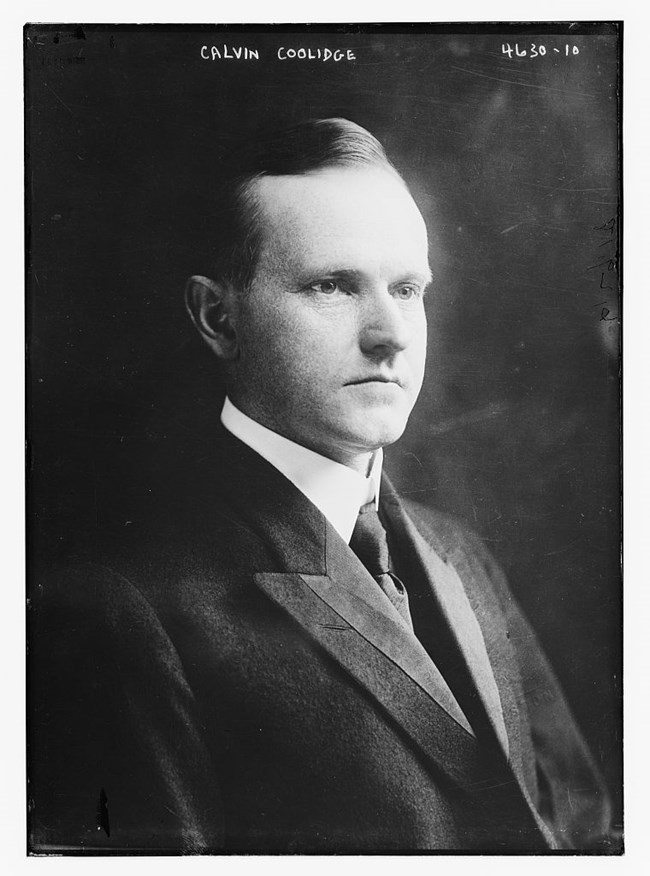 Photograph of Calvin Coolidge. He is clean shaven, facing right, and wears a dark coat.