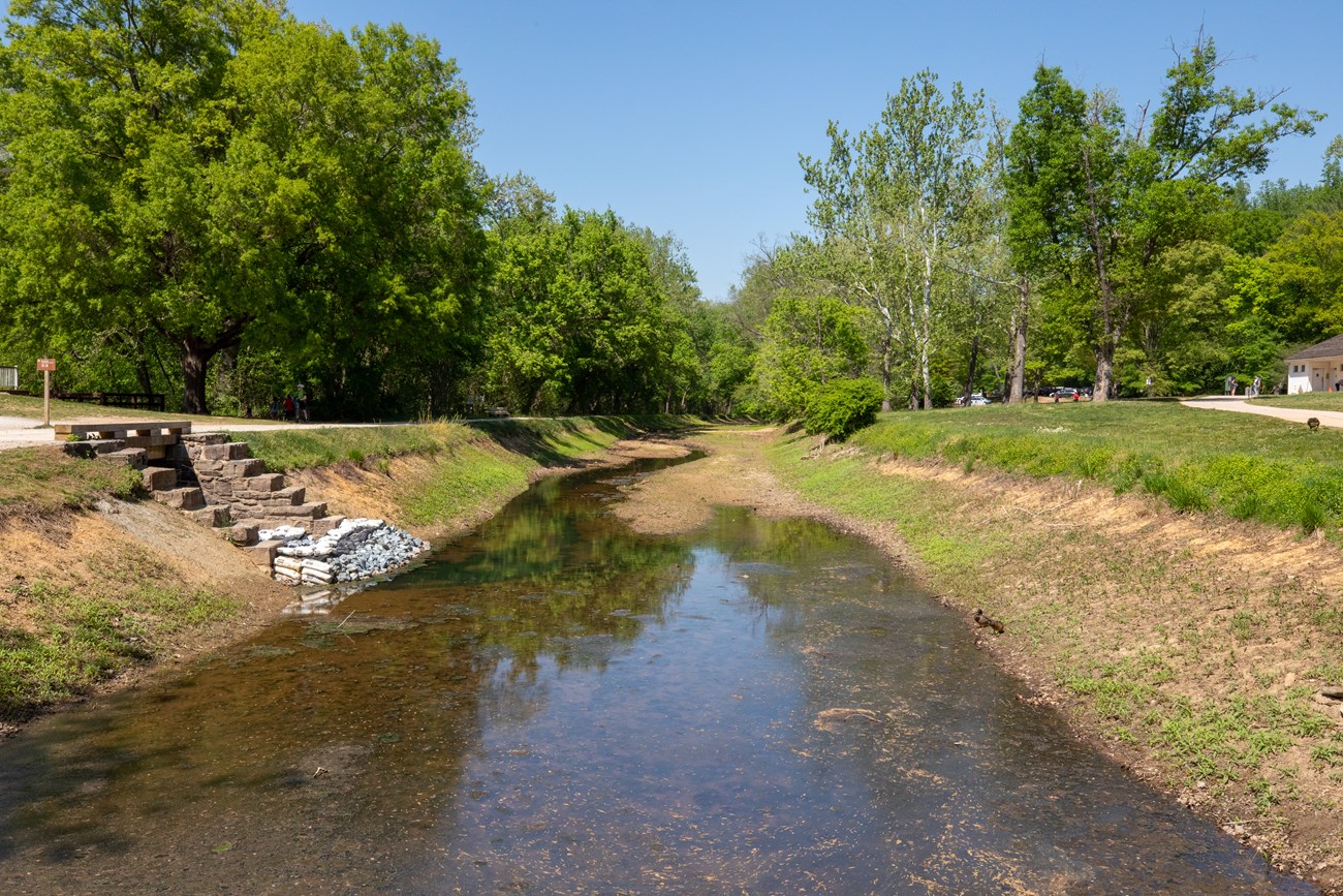 Great Falls Canal Prism A partially full of water canal prism that extends away from view. The towpath is located to the left of the canal with a paved sidewalk on the right.