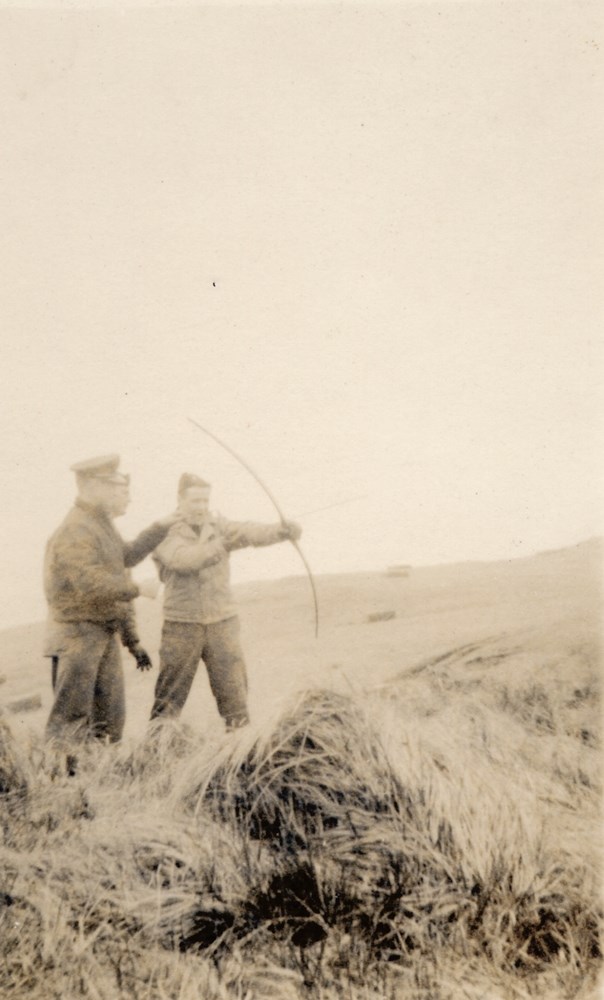 Black and white photo of three men on a grassy hill with a bow.