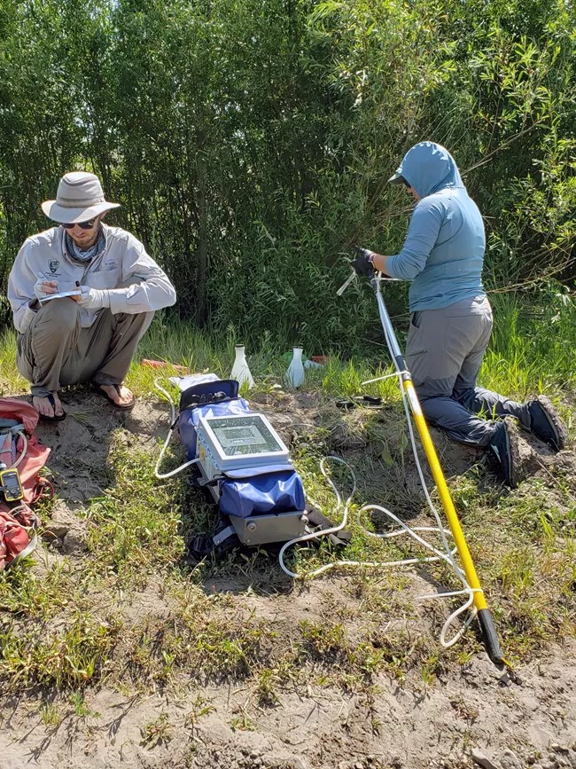 Two field workers sampling eDNA using large backpacks, sitting alongside shrubbery and a beach shore.