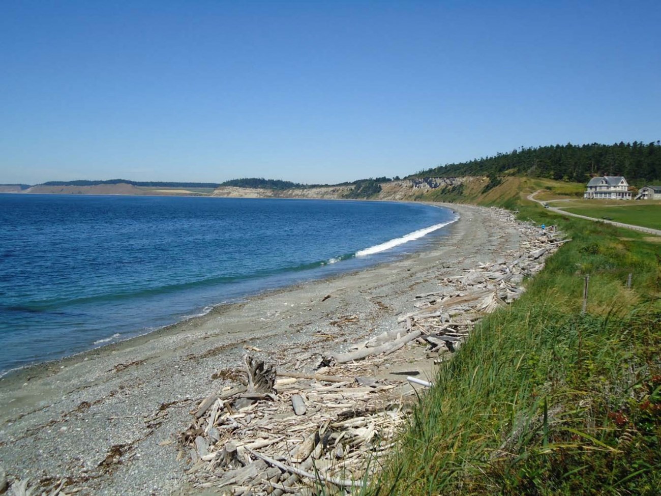 Photo of curving shoreline, pebble beach with driftwood, adjacent grassland, and distant bluffs, building, and forested hills.