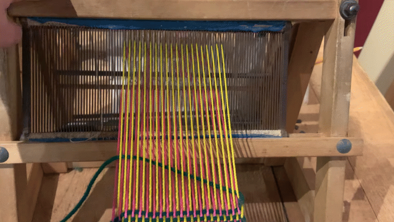 beat in gif gif of a metal comb like bar with vertical yellow and pink threads running through it is pulled forward, pushing a horizontal green yarn forward to join woven material