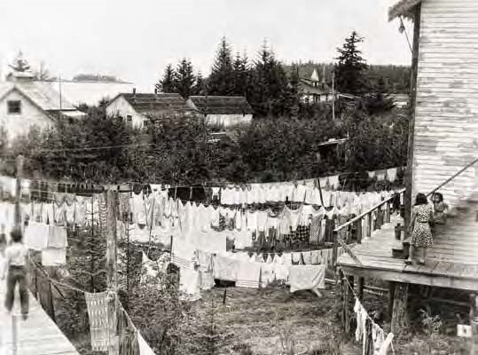 Black and white photograph of many wooden buildings with lots of laundry hanging in one yard.