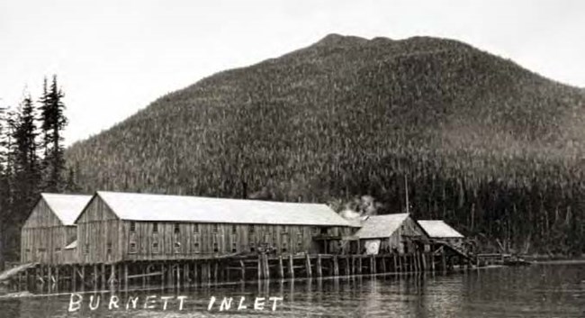 lt= Black and white photo of long wood buildings on piers in front of trees