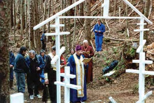 Image of a group of people including a priest viewed through a Russian Orthodox style graveyard gate.