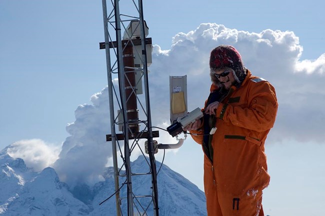 figure2_volcano monitoringweb A scientist works with instruments on a mountain top with an active volcano behind him.