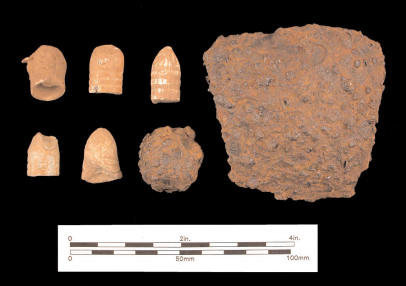 Five Fired Bullets, all dented and from impact. A rough, ball-shaped, iron case shot on middle right. A large dark brown, square-shaped shell fragment on far right.