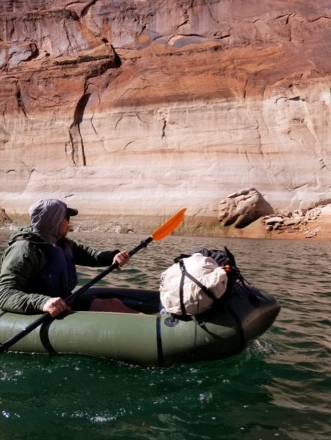 A person wearing a lifejacket paddles along a tall canyon wall in a inflatable kayak with their gear tied to the front.