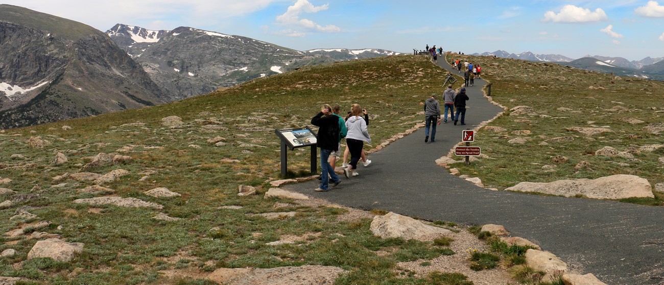 people climb paved trail with mountains in background