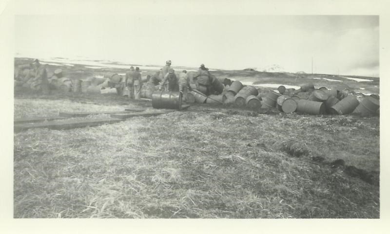 lt= Black and white photo of men rolling barrels up an incline