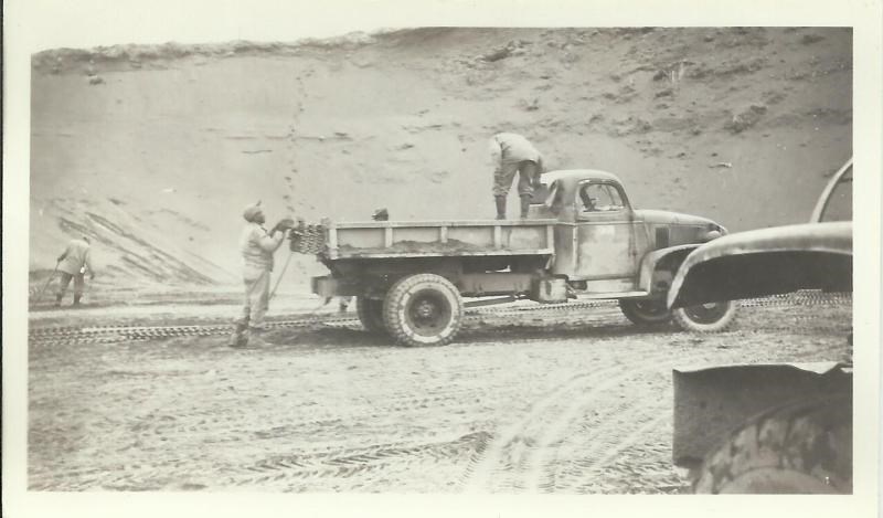 lt= Black and white photo of men unloading wire mats from a truck with an earth bank behind them.