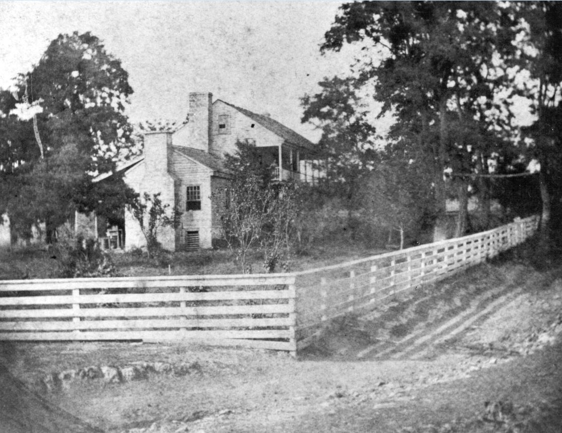 Black and white photo of large, two-story house with porch and fenced-in yard.