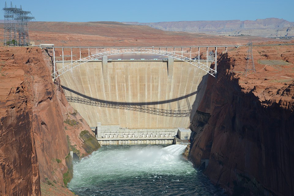 A dam with water form a river at its base and red rocky cliffs on the right and left of the dam. A bridge spans the river at the top of the cliffs. Power lines are seen at the top of the cliff on the left.
