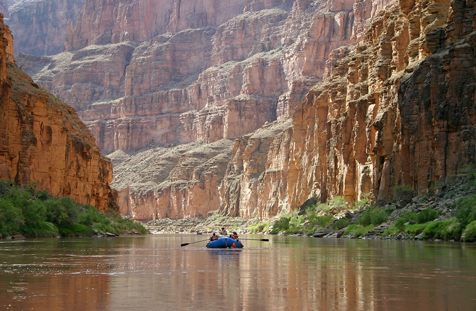 A blue raft in the middle of a river with towering red and orange cliffs on both sides of the river. Green vegetation is at the base of the cliffs by the river