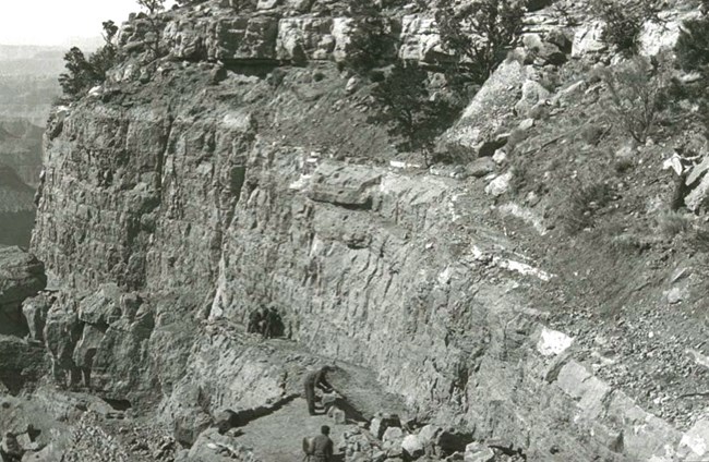 Edge of a cliff, showing a ledge that leads down into the canyon. Workers using pickaxes near the entrance.