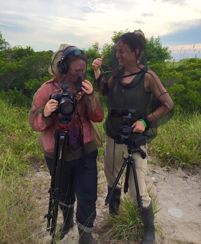 lt= Two women stand in a marsh with camera eqipment