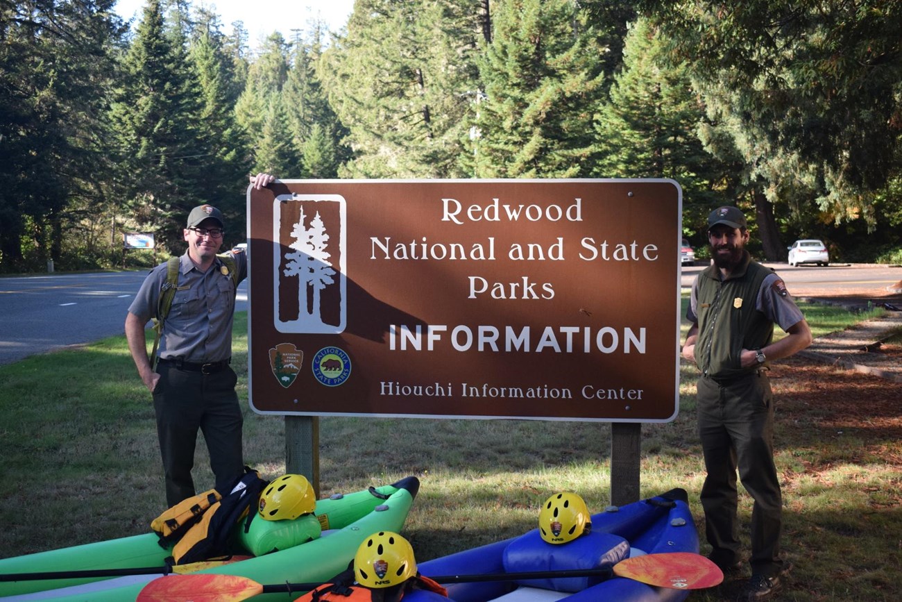 Redwoods National and State Parks two park rangers standing by the Redwoods National and State Parks sign with two kayaks in front.