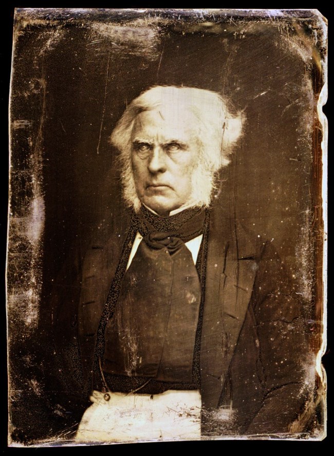 Photo portrait of man with white hair and stern expression wearing dark clothing