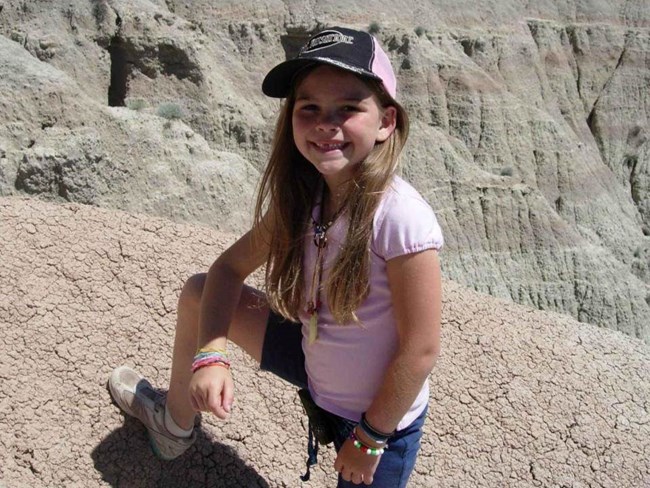 Young girl in hiking attire standing amid badlands formations on a sunny day.