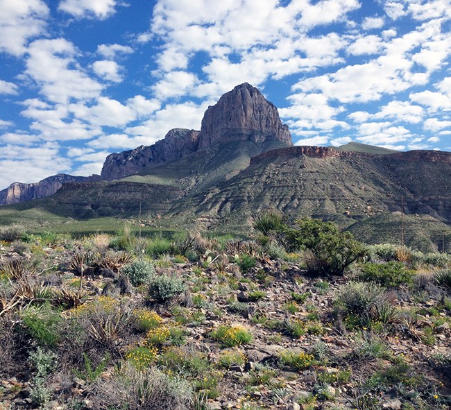 Tall rocky mountains surrounded by desert with scattered clouds above