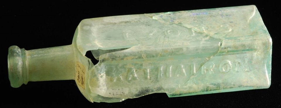 Greenish glass bottle, broken and pieced together, with raised lettering.