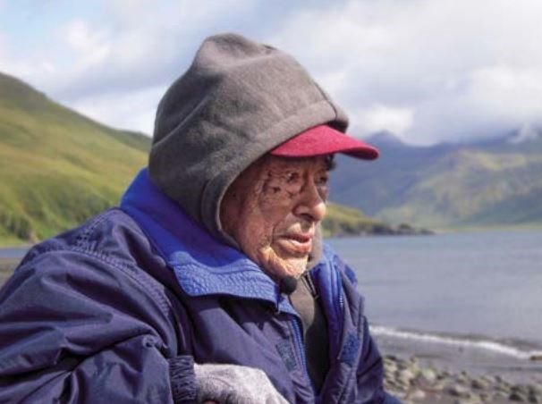Color photo of a man in a blue raincoat looking out from shore.
