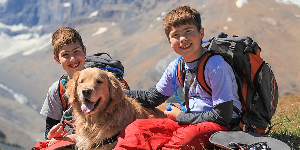 Two kids wearing backpacks sit with a leashed golden retriever