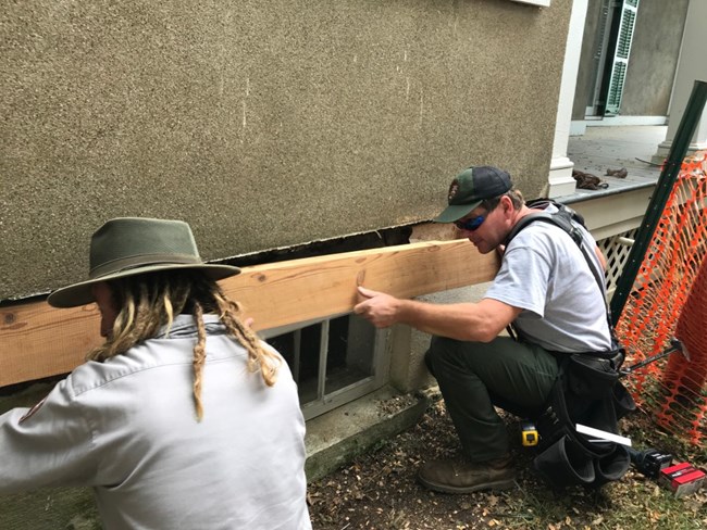 Two men apply a fresh board to the base of a building.