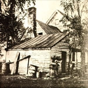 Black and white photograph of multi-part white wooden house. There are barrels and wood planks next to the house and two young African American boys stand in the front.