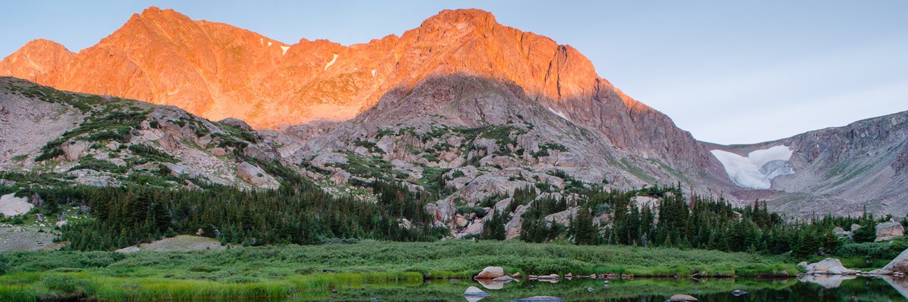 Mountains with an orange glow from the sun with trees and a mountain lake in the foreground