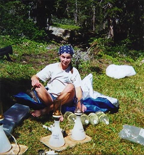 A woman sits on the ground in the woods with canisters of water
