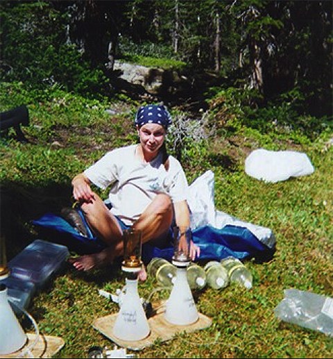lt= A woman sits on the ground in the woods with canisters of water