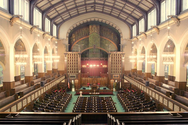 Balcony view of rows of brown pews, green carpet, and tan walls with arches.