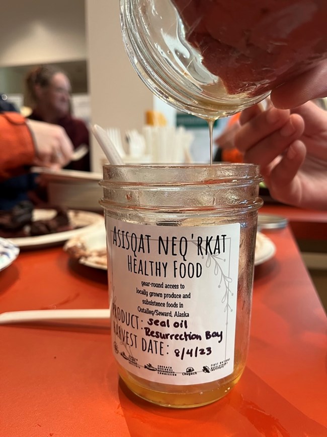 Oil is poured into a jar of seal oil that reads "ASSQAT NEQ'RKAT HEALTHY FOOD" and indicates it was harvested from Resurrection Bay on 8/4/23