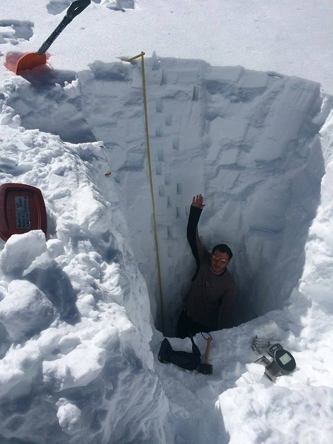 USGS snow chemistry sampling man waves from 10 feet down inside a hole dug into deep snow, with red snow shovel standing upright on the snow surface above