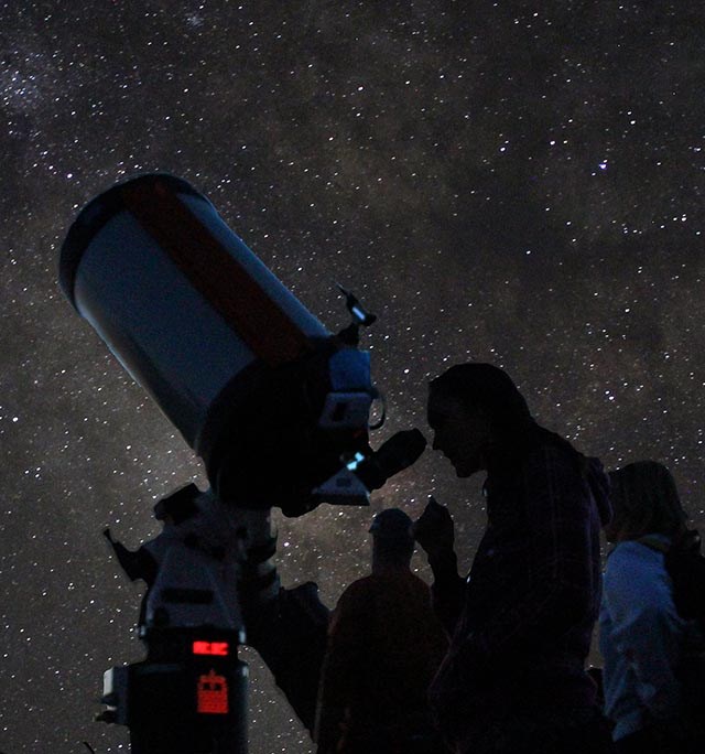 under a starry night sky, several people are taking turns looking through a large telescope.